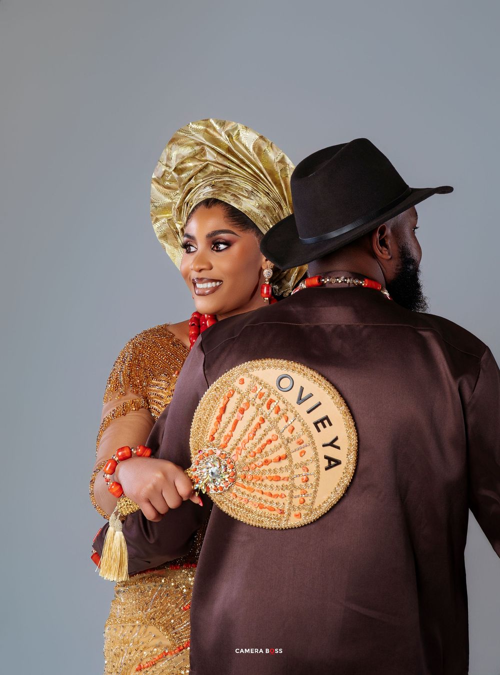 Couple in traditional African attire with gold headwrap and brown jacket featuring circular Ovita emblem posing back-to-back.