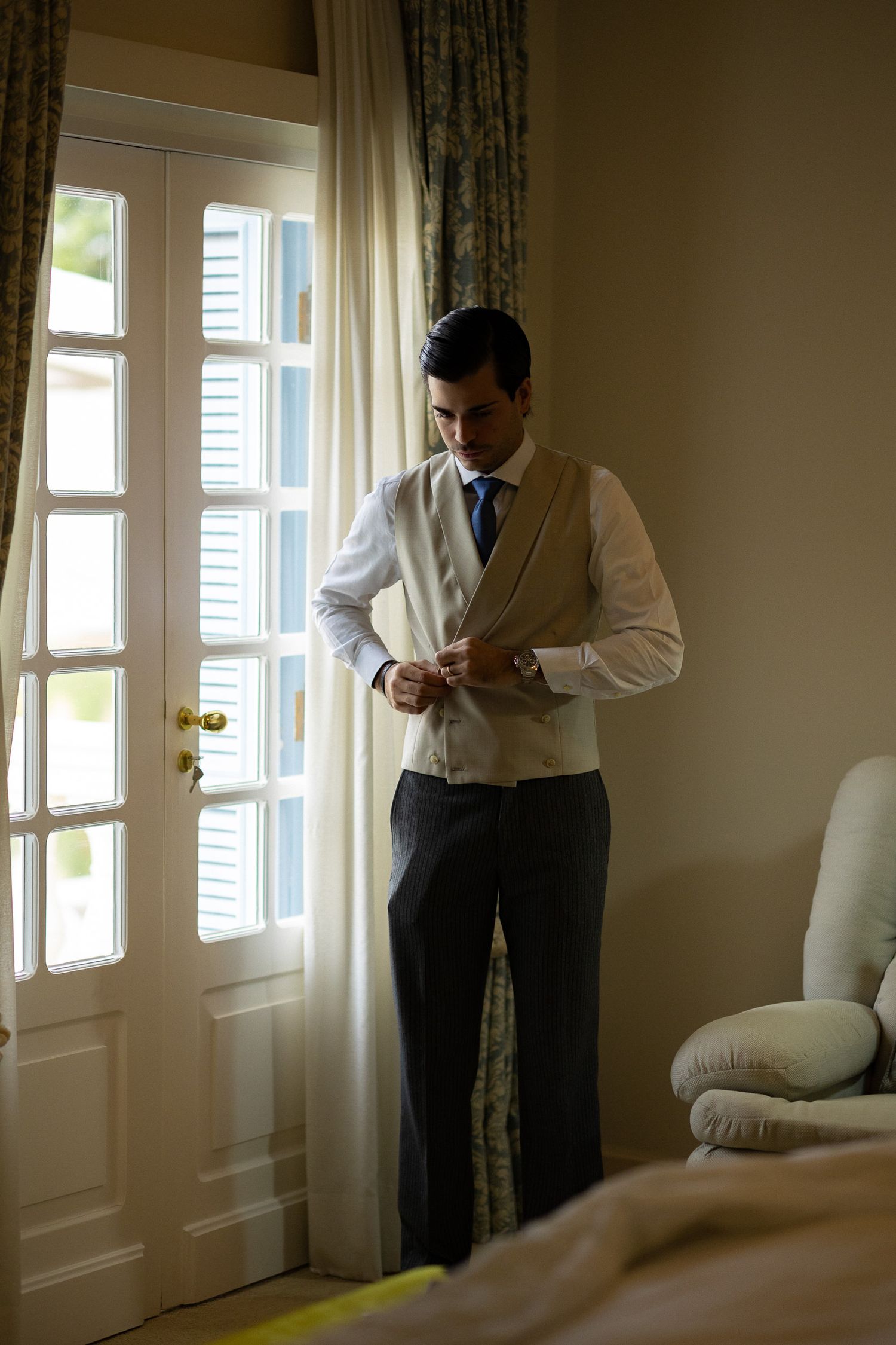 Man in formal business attire adjusts his tie while standing in a sunlit room with white curtains.