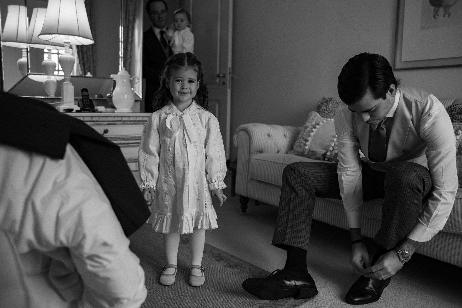 A black and white photograph shows a small child in a dress standing near an adult seated on a couch.