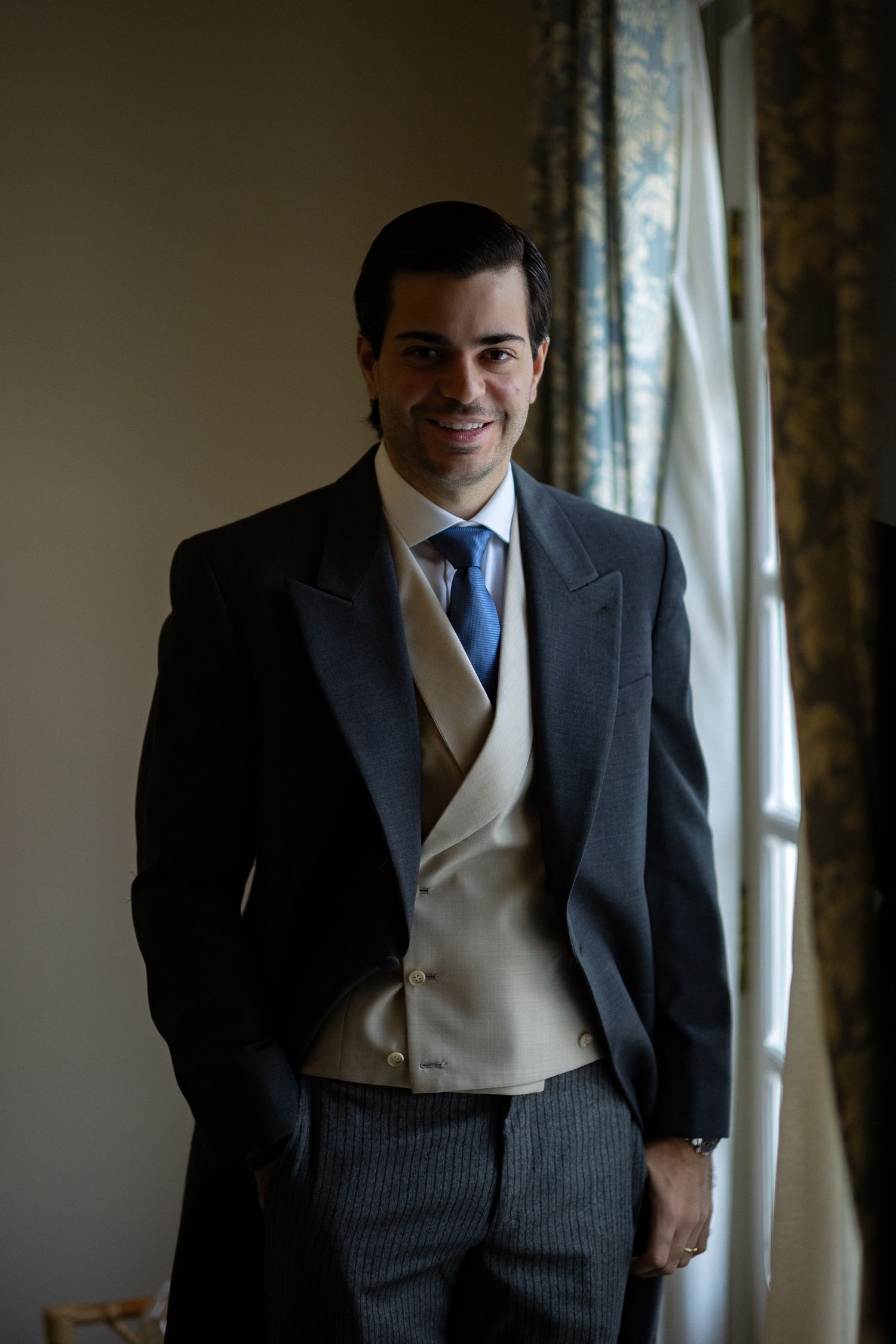 A person wearing a formal dark suit with a cream vest stands by a window in elegant attire.