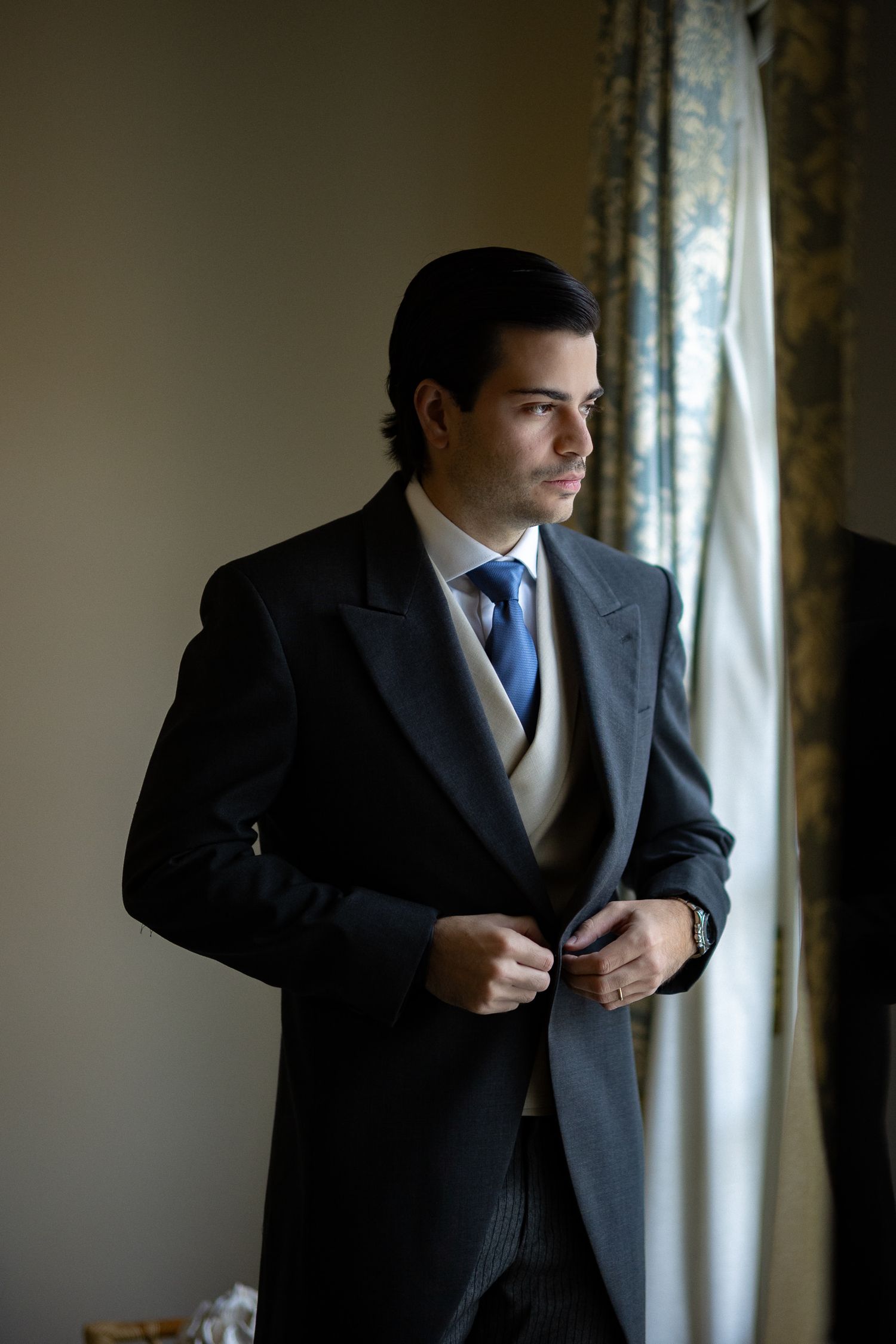 A person adjusts their dark suit jacket while standing near a window with natural lighting.