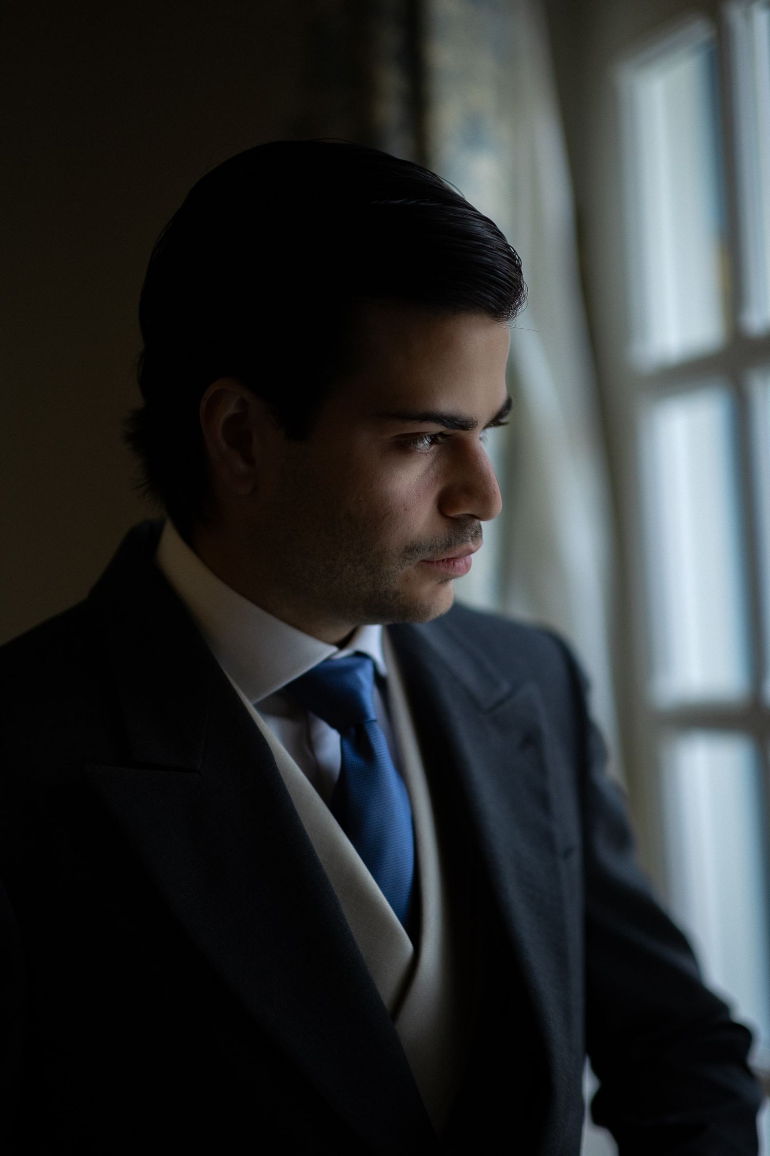 A businessman in a dark suit and blue tie gazes contemplatively through a window in moody lighting.