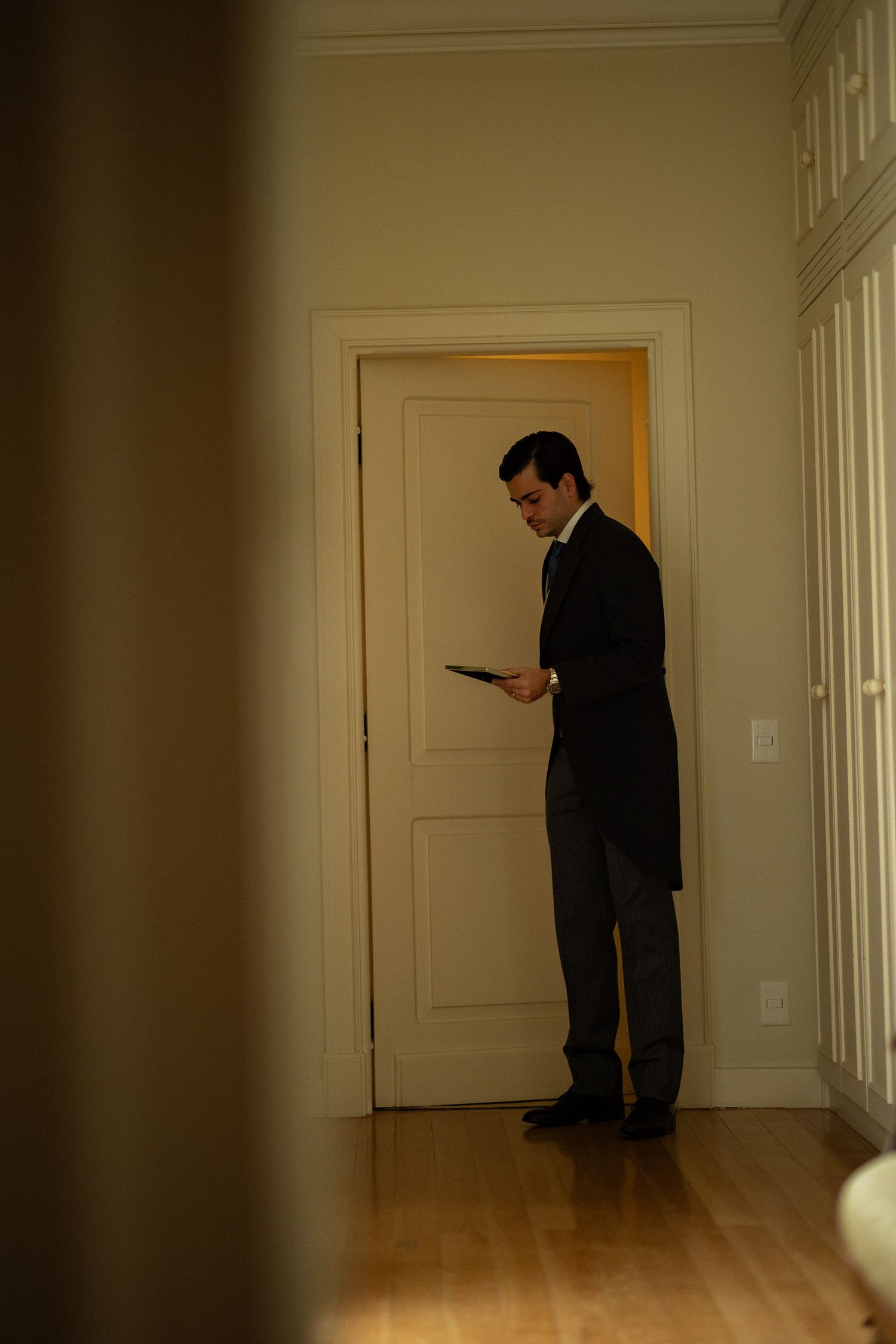 A person in a dark suit stands reading in an empty white hallway with hardwood floors and natural lighting.