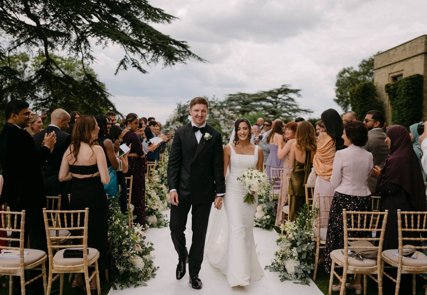 Outdoor wedding ceremony setup with white chairs and floral arrangements under cloudy sky.