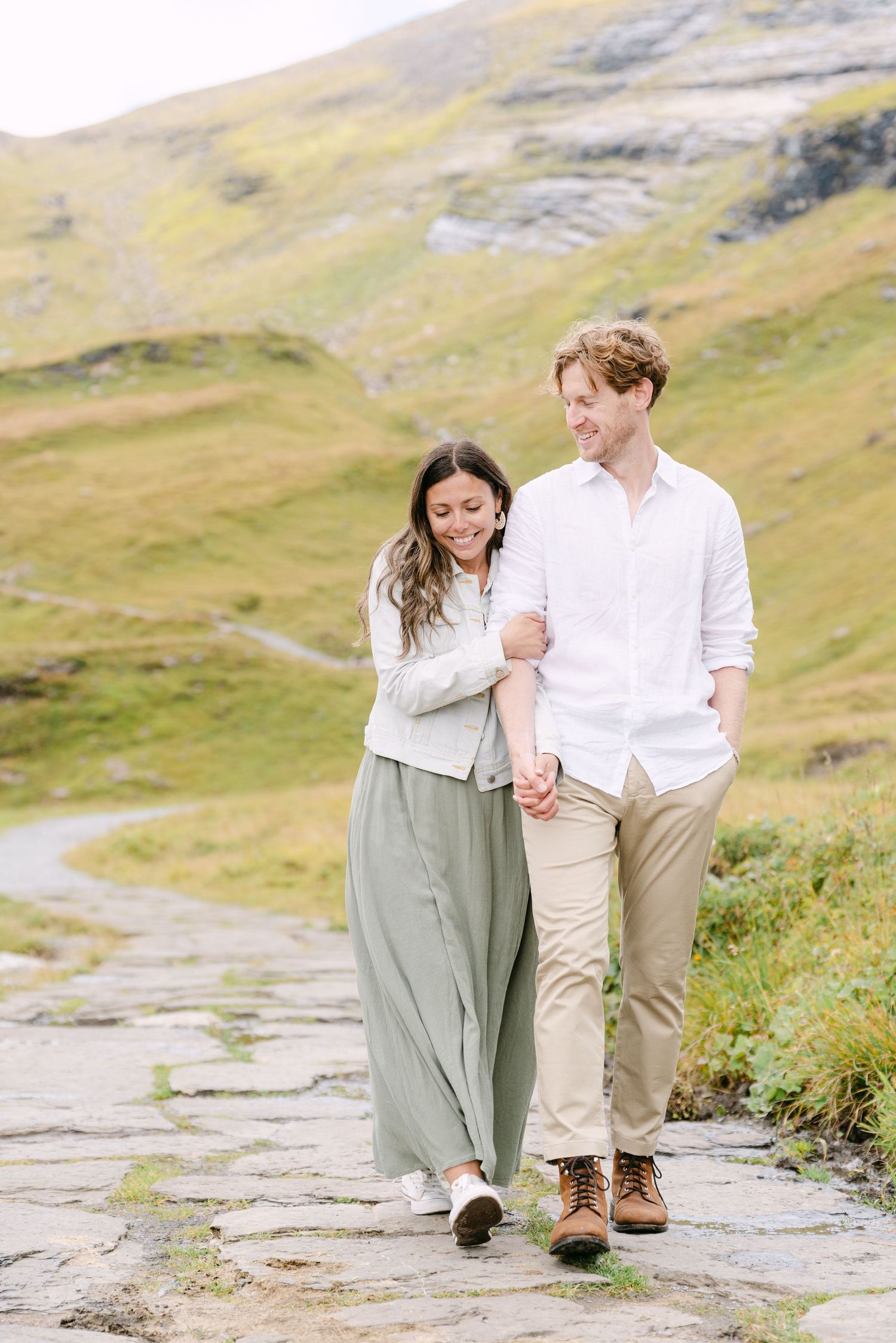 A couple walks together on a mountain path surrounded by green hills and rugged terrain.