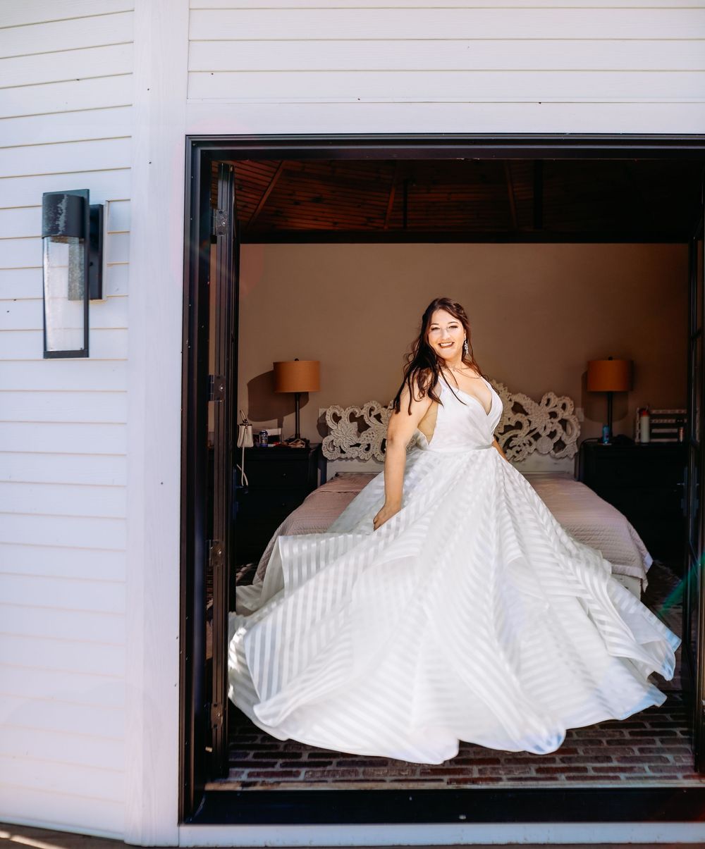 A bride in a stunning white striped ballgown twirls gracefully in a doorway with warm lighting and bedside lamps visible.