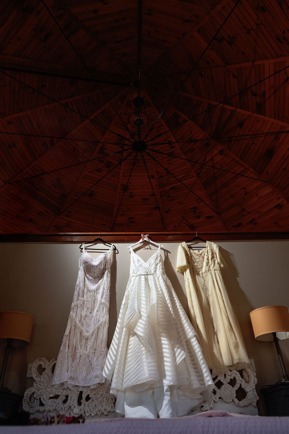Multiple vintage white lace dresses hanging in a row against a dark wooden ceiling.