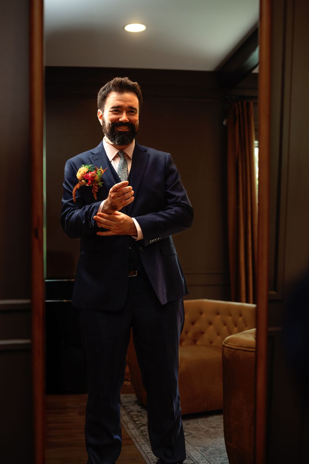 A person in a navy blue suit stands in a warmly lit room with wooden columns and a brown leather couch.