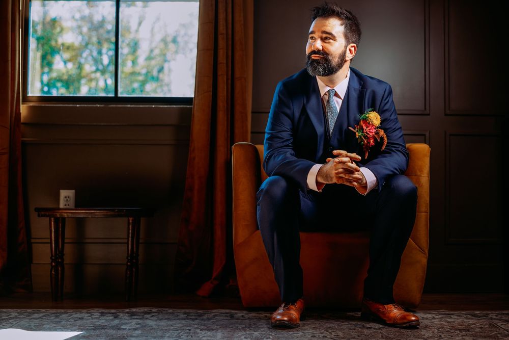 A groom in a navy suit and brown shoes sits thoughtfully on a leather chair near a window with an orange boutonniere.