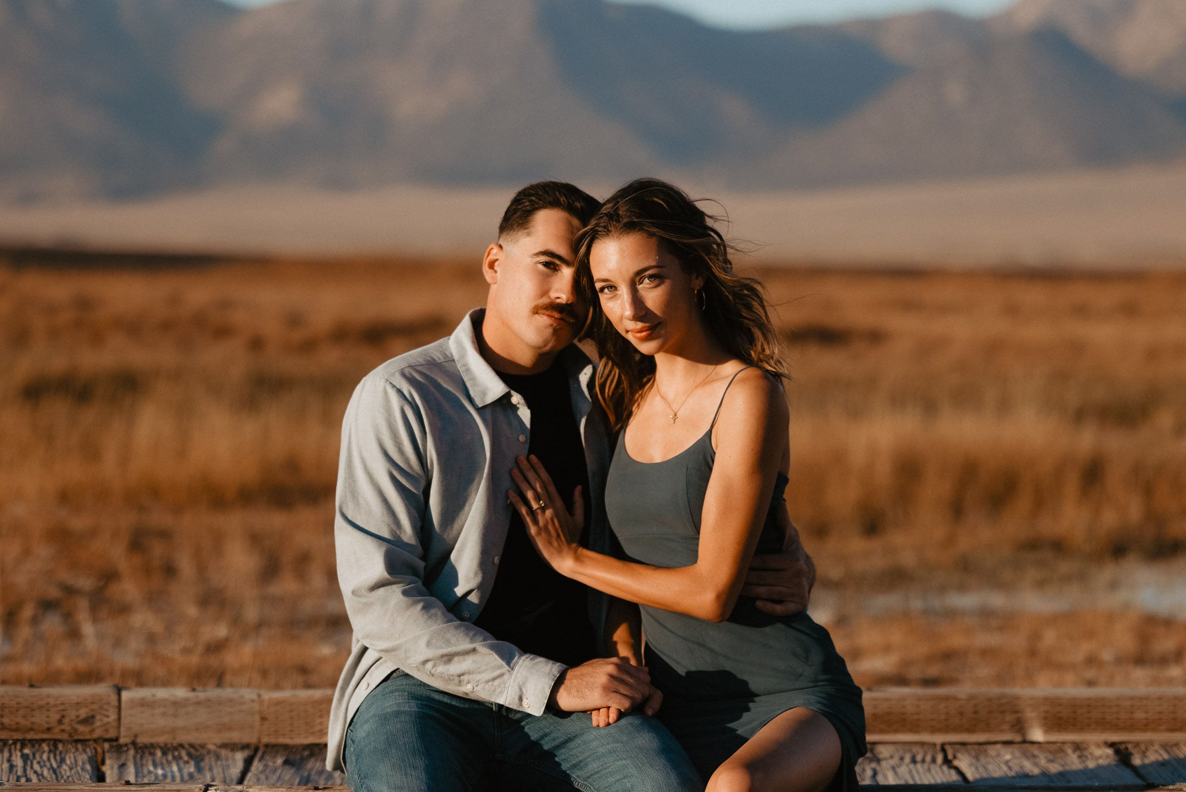 Romantic couple embraces in a golden field during sunset with mountains in the background.