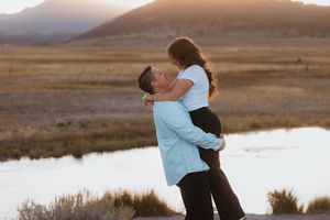 Couple poses together near a serene lake with mountains at sunset.