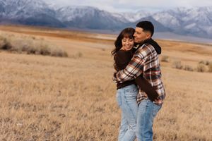 Playful couple poses in casual outfits against mountain landscape backdrop.