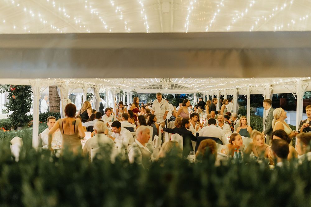 A panoramic view of wedding guests seated in white chairs during an outdoor ceremony at sunset.