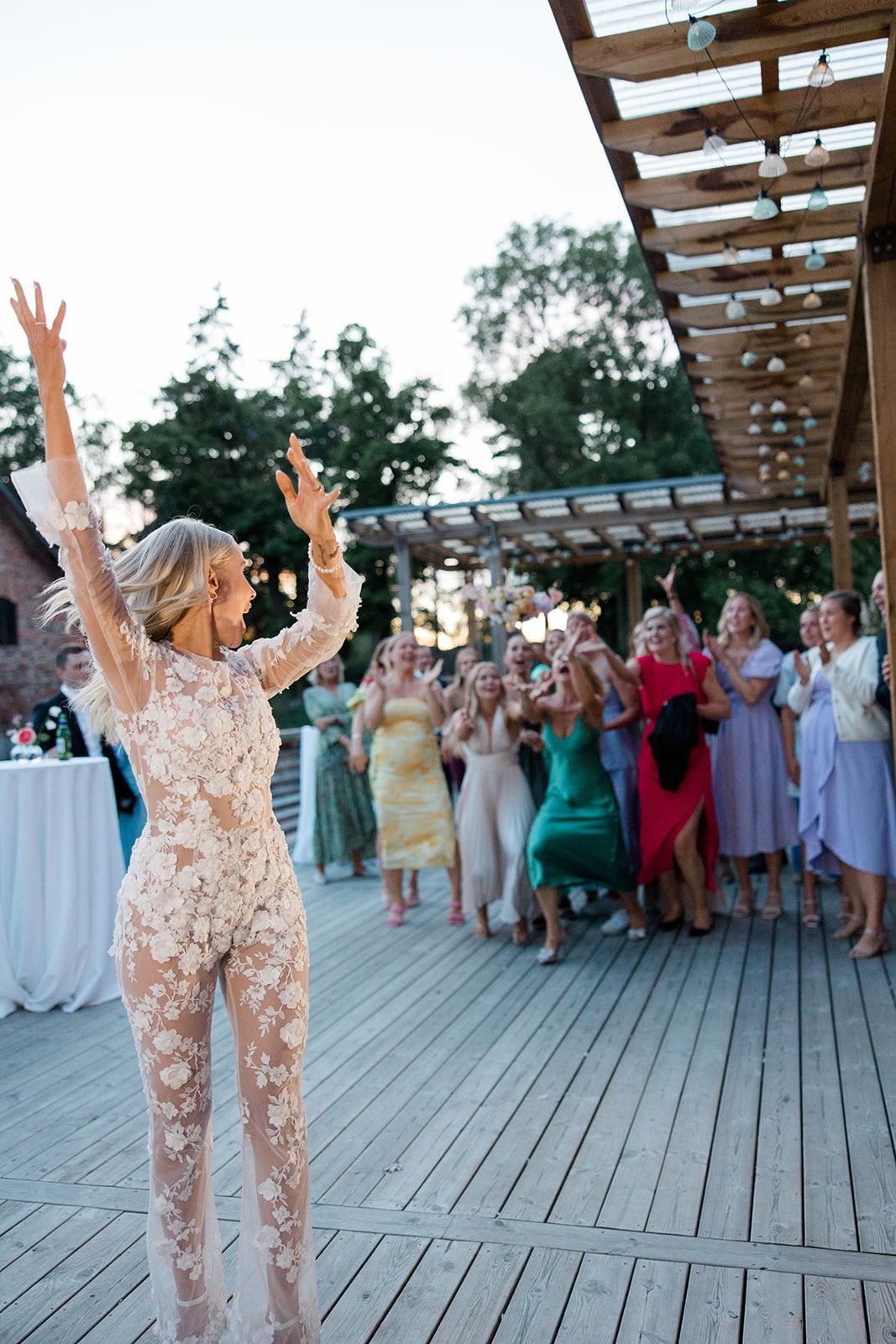 Wedding guests dance and celebrate on an outdoor wooden deck under a pergola during sunset reception.
