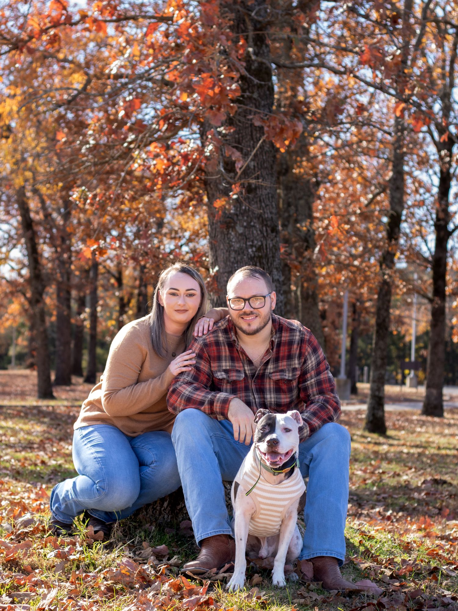Husband and wife smiling with their dog in the fall leaves.