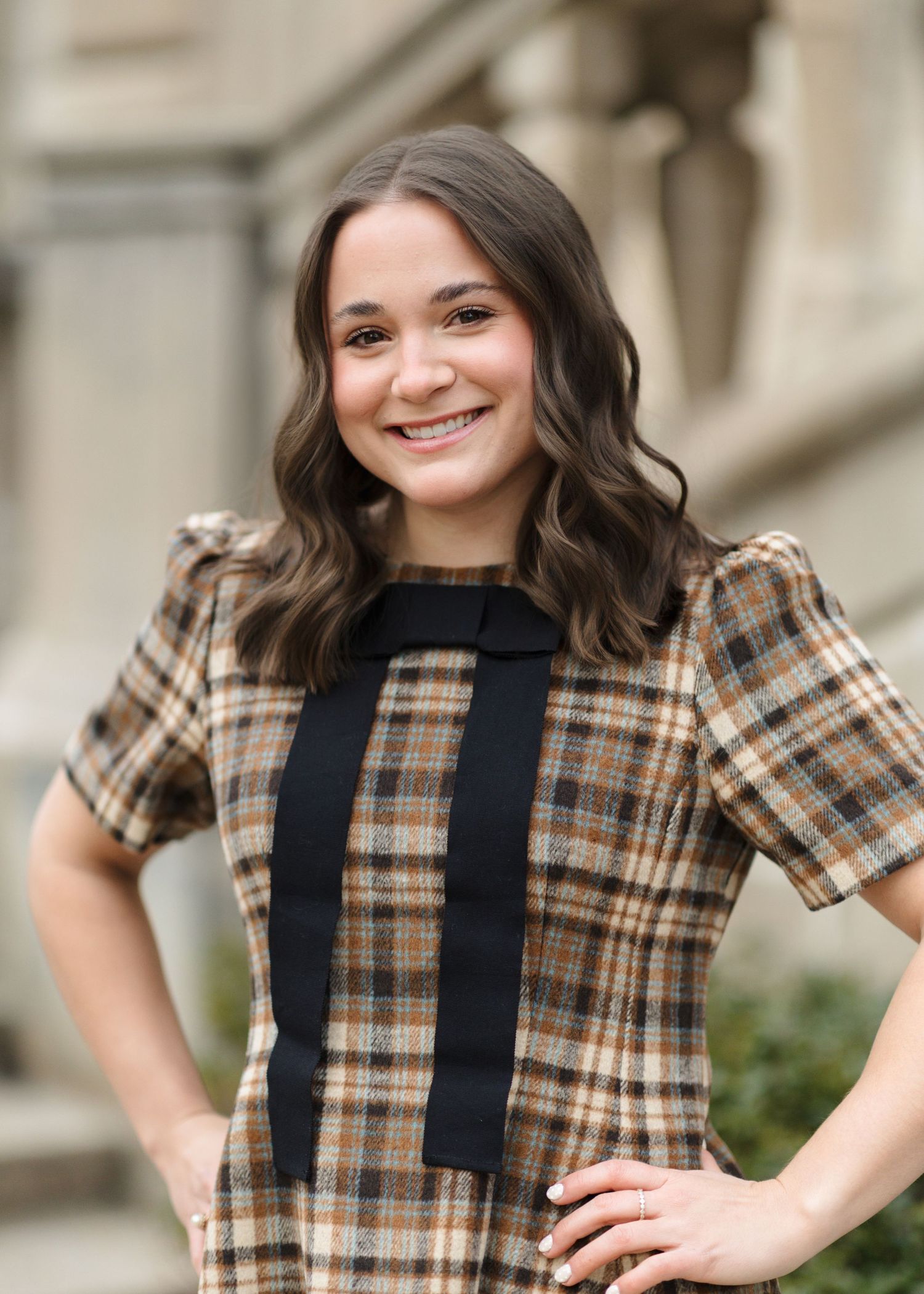 A woman in a brown plaid short-sleeve dress with black vertical stripes poses outdoors with a warm smile.