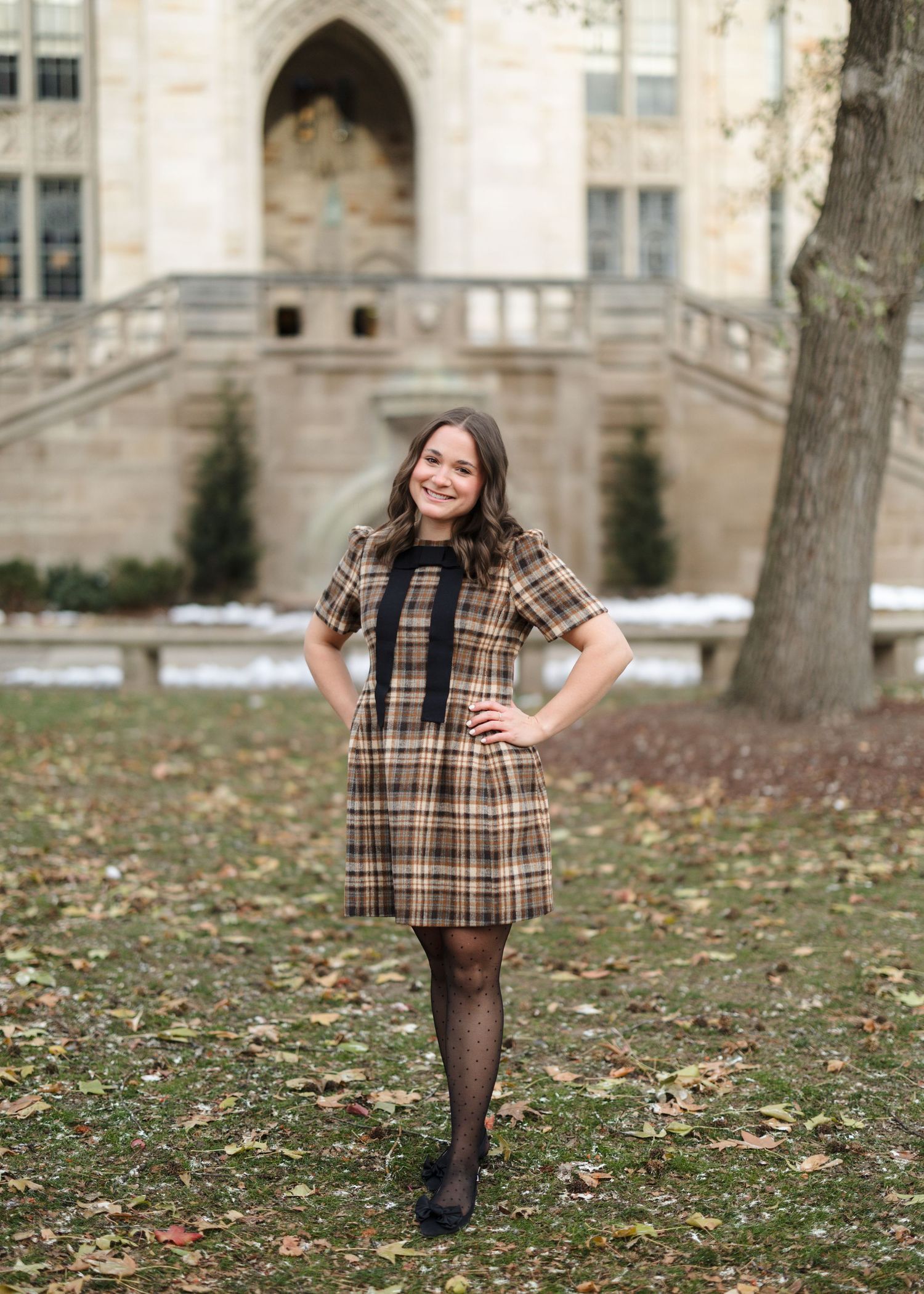 A person in a plaid dress and tights poses outdoors on a grassy lawn with autumn leaves and stone buildings in background.
