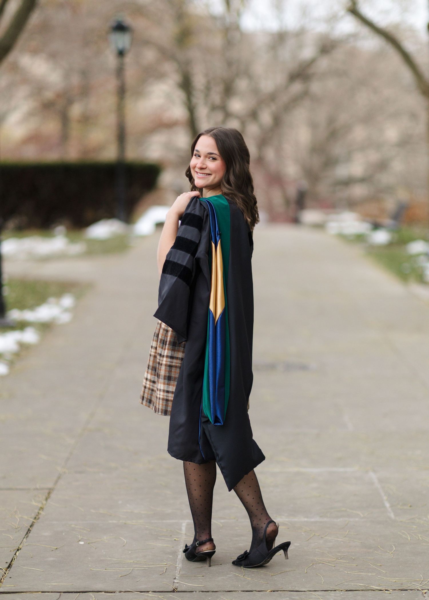 A person wearing a dark coat and plaid skirt walks along a snowy path while carrying a colorful scarf.