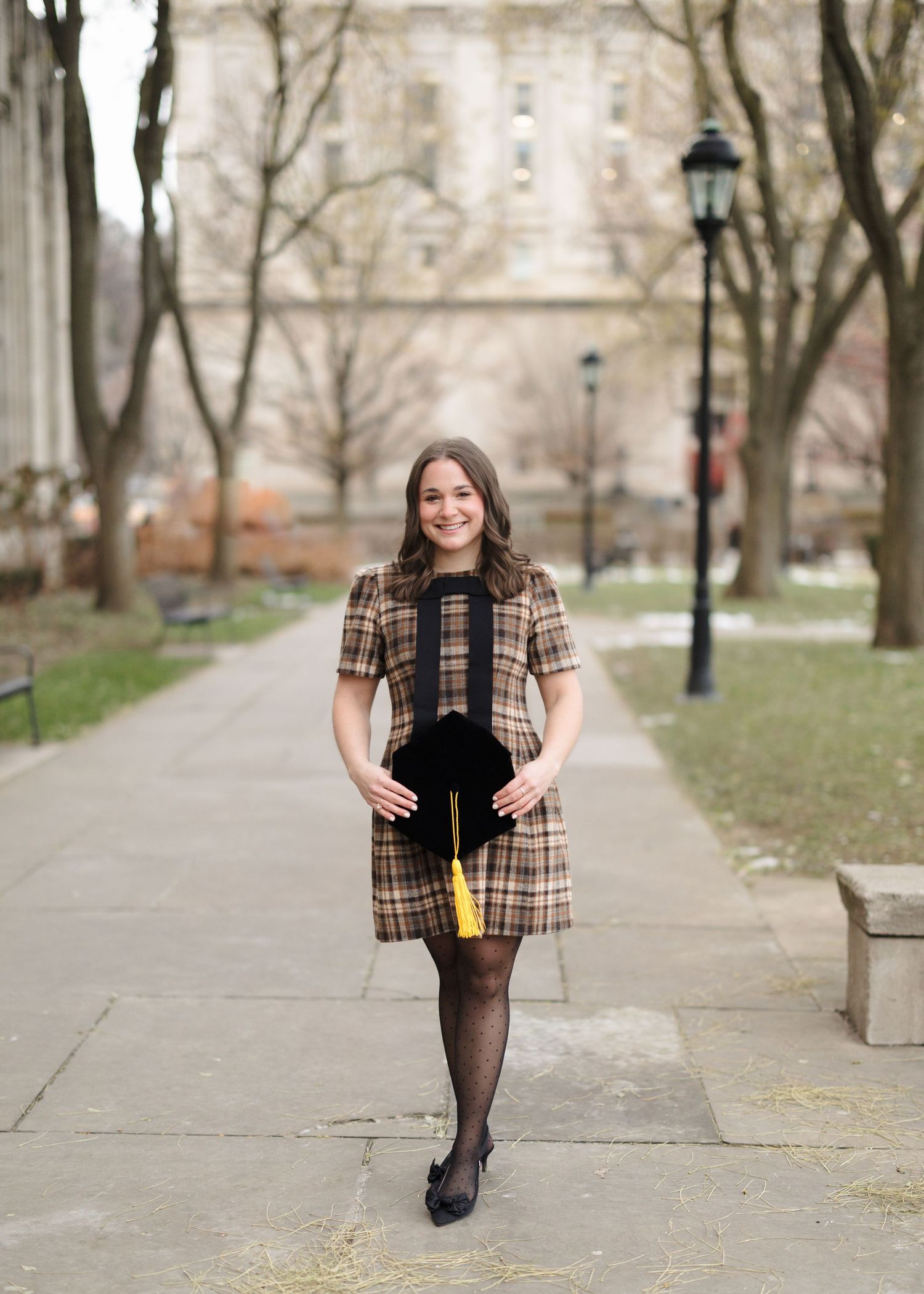 A person in a plaid dress and black tights stands on a tree-lined pathway with lamp posts and buildings in the background.