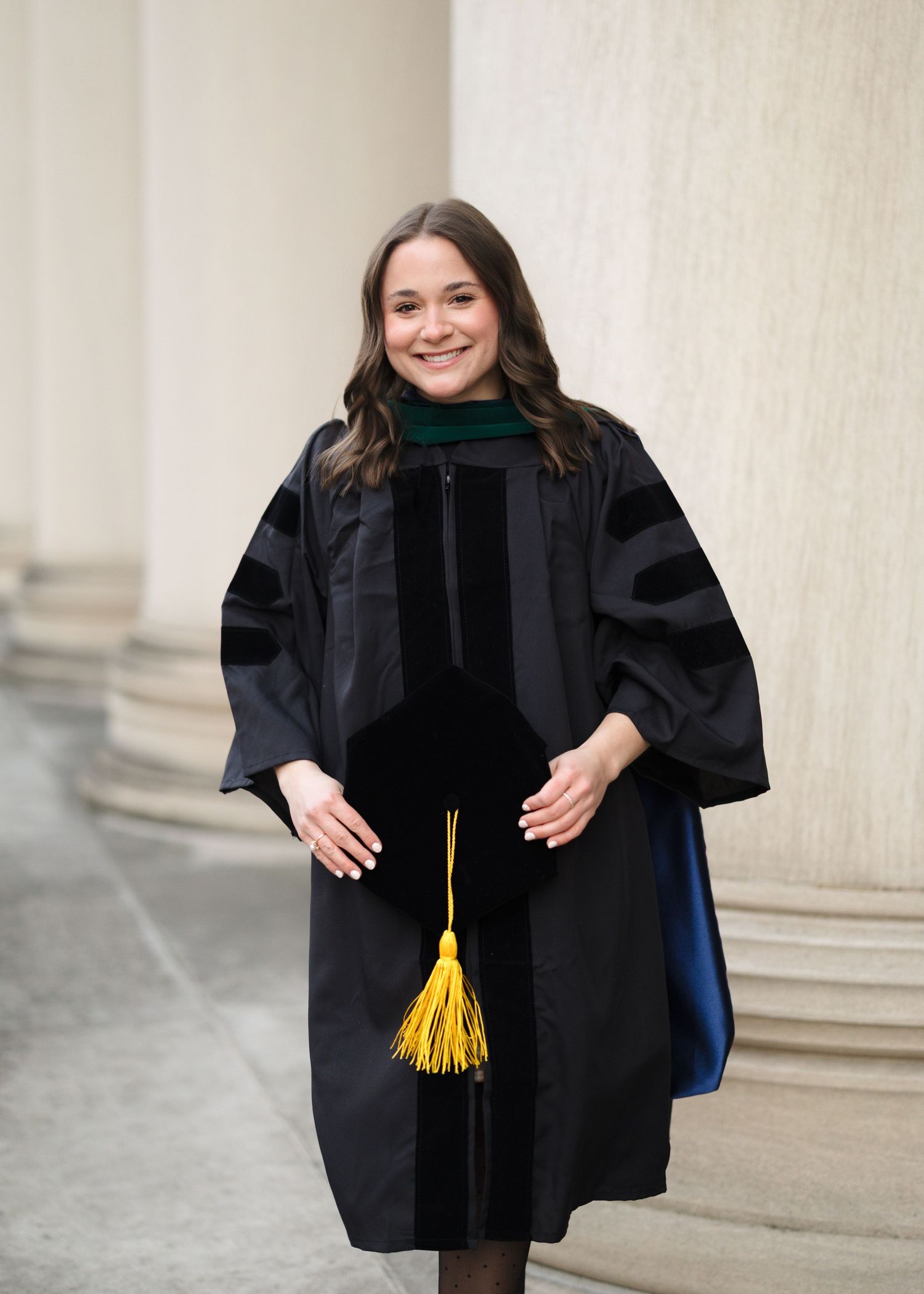 Graduate wearing black academic regalia with gold tassel poses in front of stone columns during commencement ceremony.