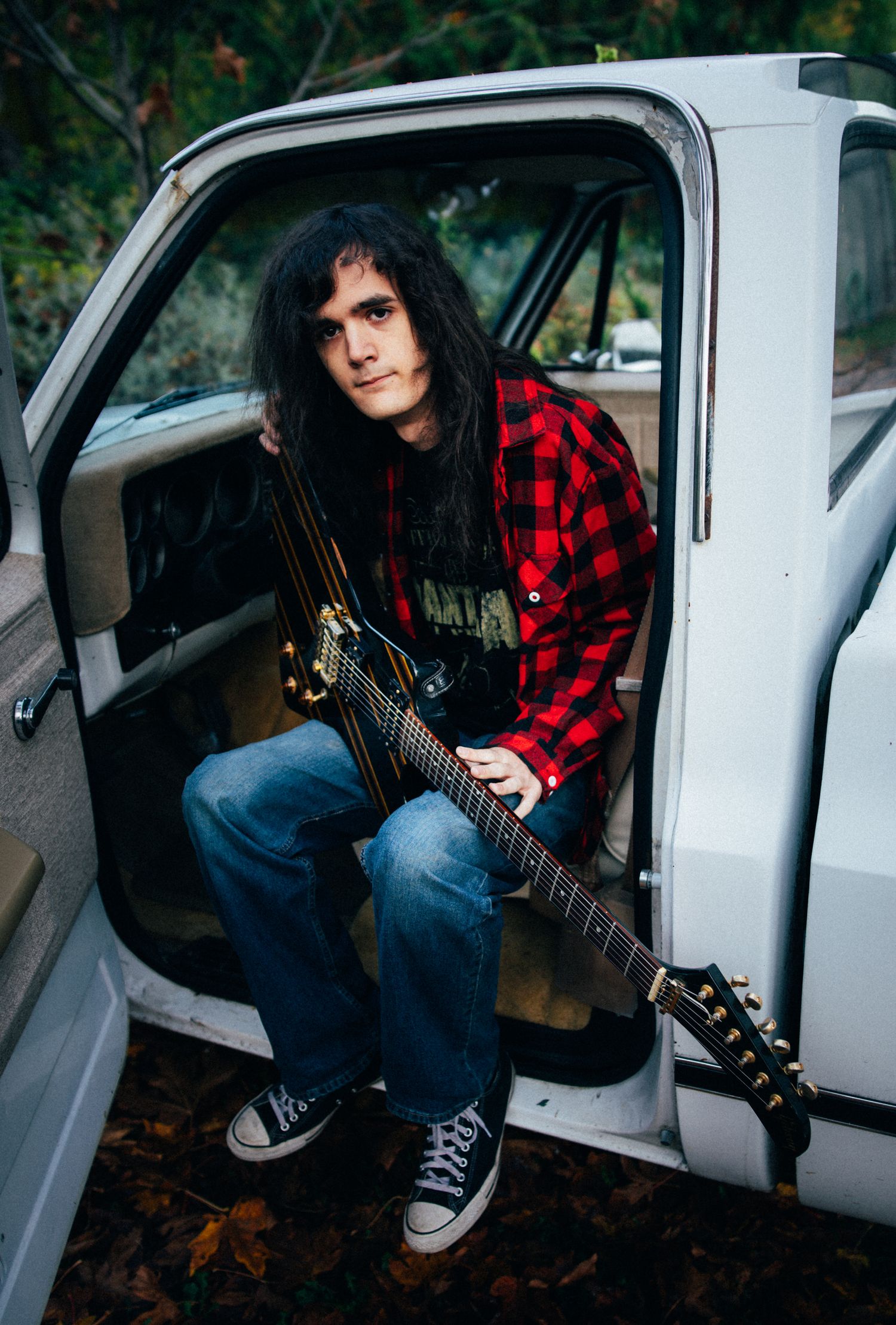 Young male guitarist with long dark hair sits in truck door holding electric guitar outdoors.
