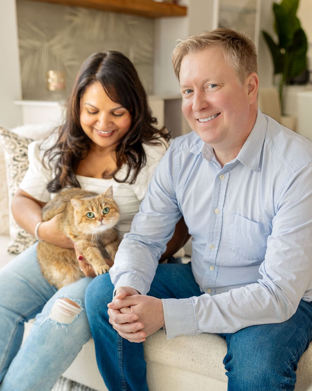 A couple sits together on a couch while playing with their ginger cat in a bright, cozy living room setting.
