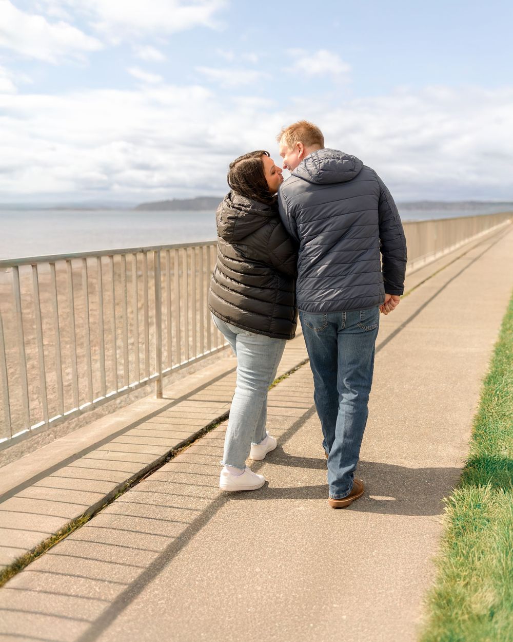 Two people in puffy jackets walking together along a coastal promenade with a metal railing and ocean view.