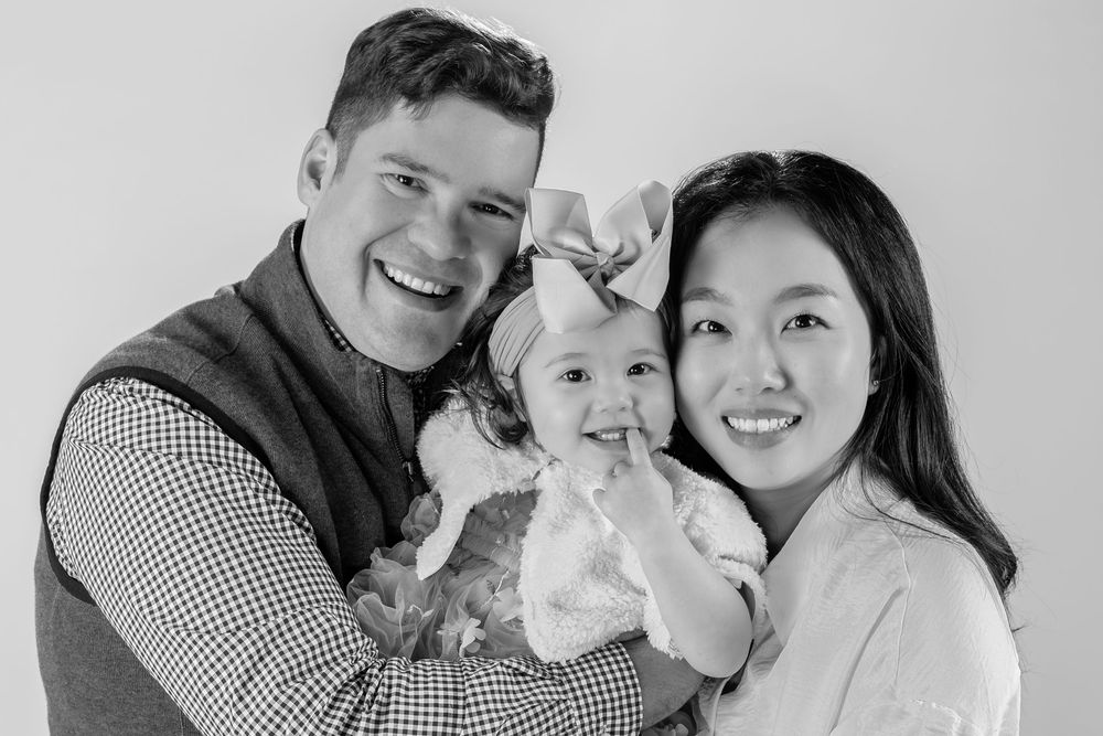 Black and white family portrait of smiling parents holding their baby girl with a large bow in a studio.