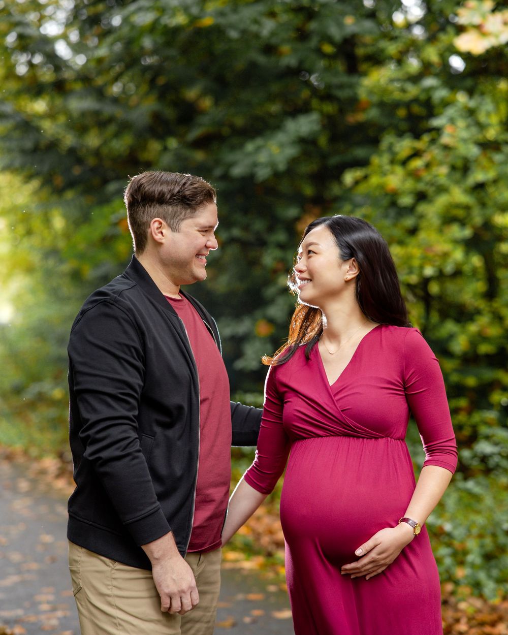 A couple in a burgundy shirt and red dress share joyful moments during an outdoor maternity photoshoot.