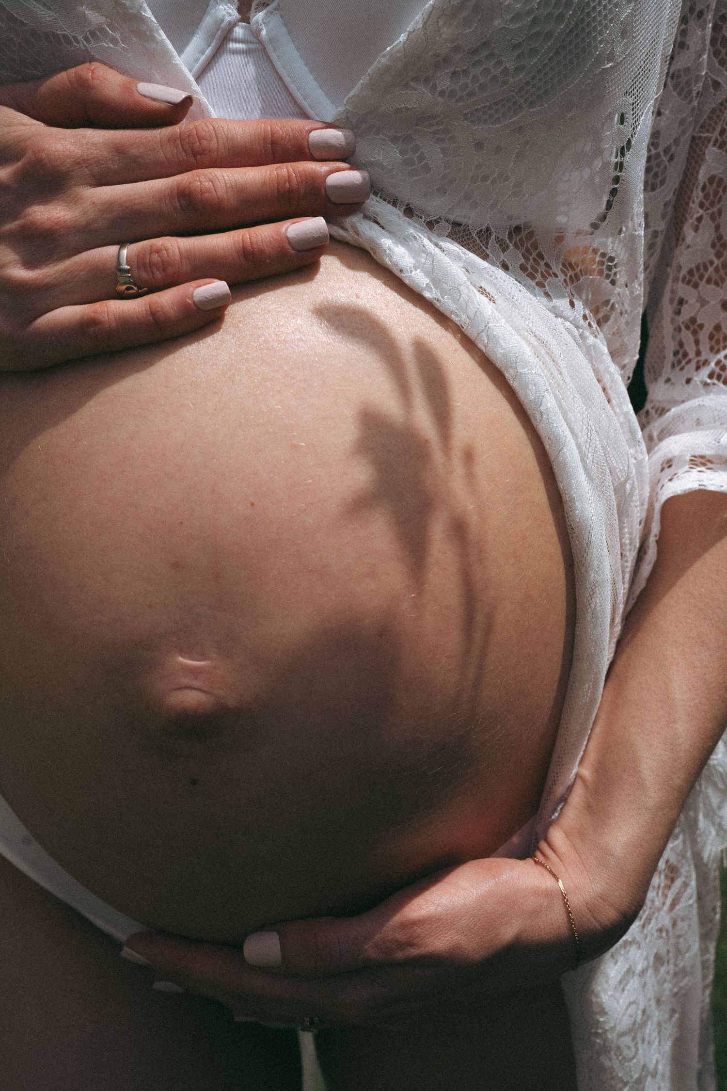Close-up of hands cradling pregnant belly in white lace dress.