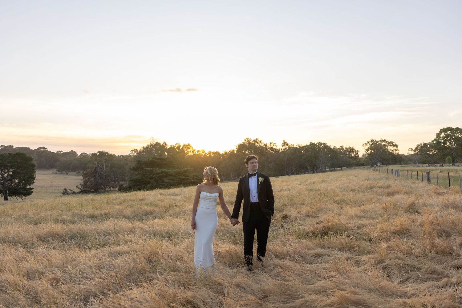 A couple in wedding attire walks through a golden field at sunset with trees silhouetted in the background.