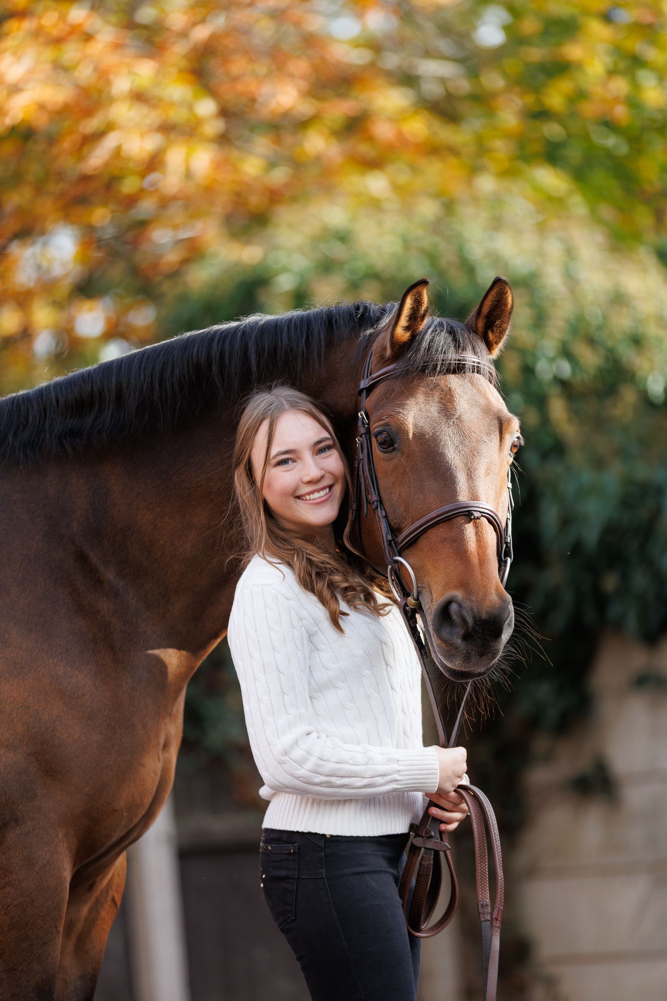 A brown horse and rider in a white sweater pose together against autumn foliage in soft natural lighting.