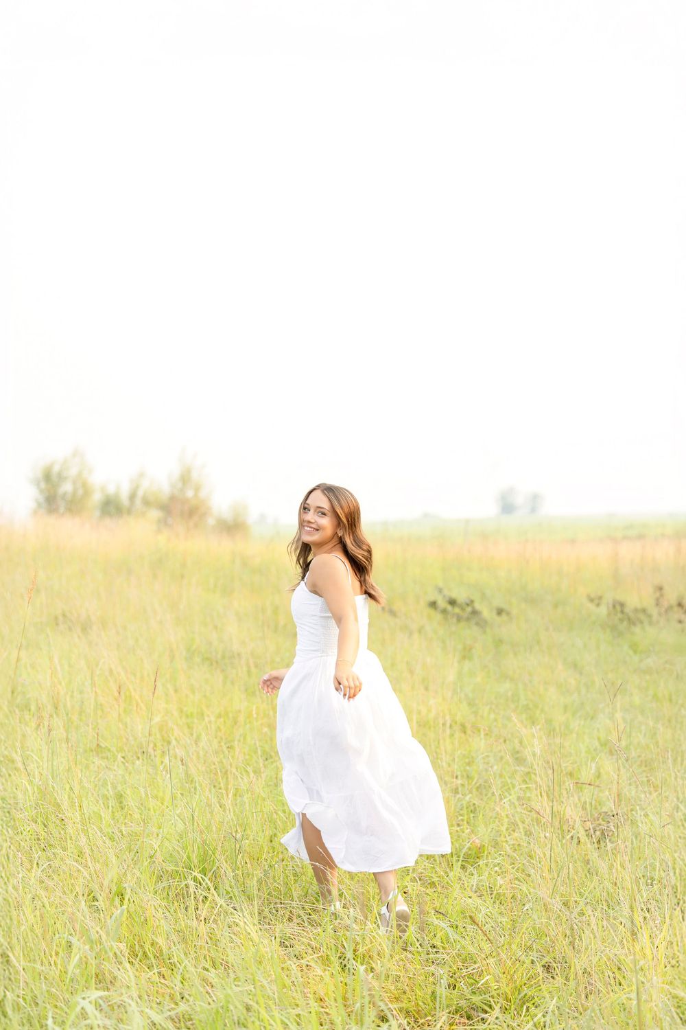 A high school senior girl in a flowing white dress twirls gracefully through a sunlit meadow of tall grass during photoshoot Clear Lake Iowa