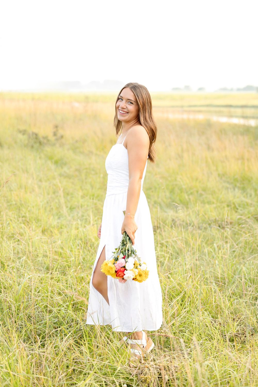 A person in a flowing white dress holds colorful flowers while standing in a sunlit meadow.