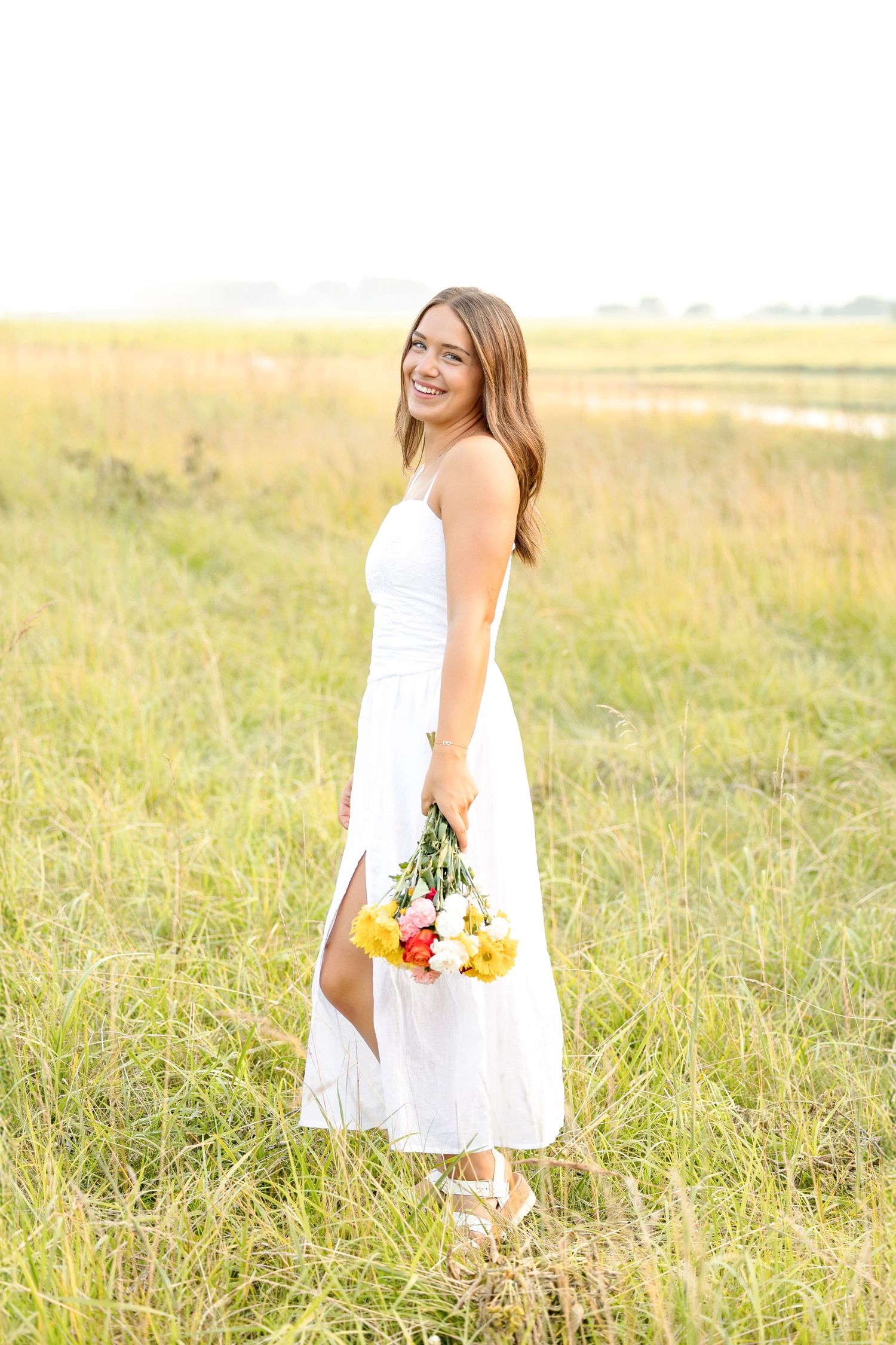 A person in a flowing white dress holds colorful flowers while standing in a sunlit meadow.