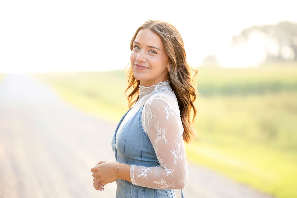A senior in a denim dress poses outdoors during golden hour with soft sunlit glow in the background on Iowa country roads.