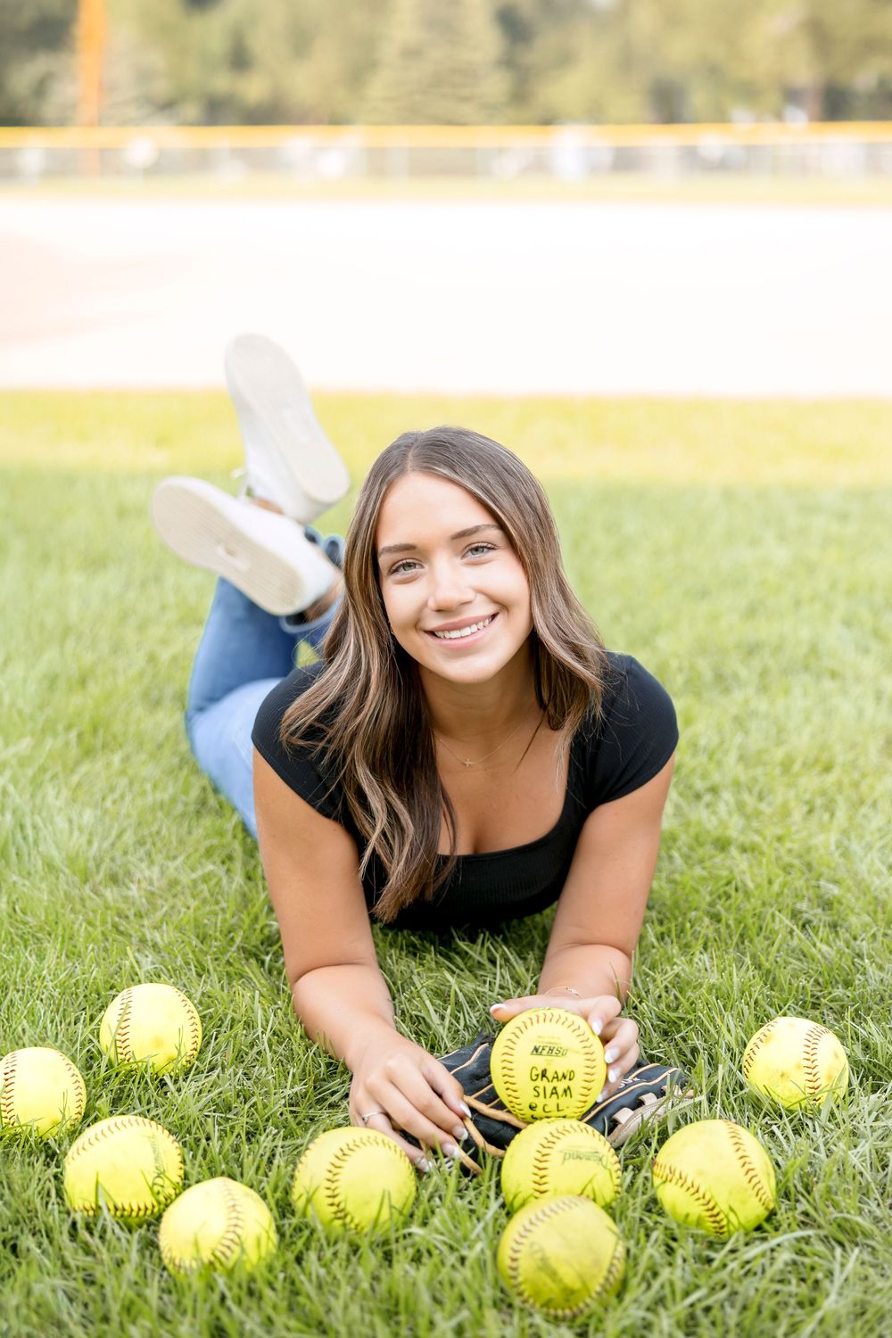 A person lies on grass with yellow softballs arranged in front of them during an outdoor photo session.