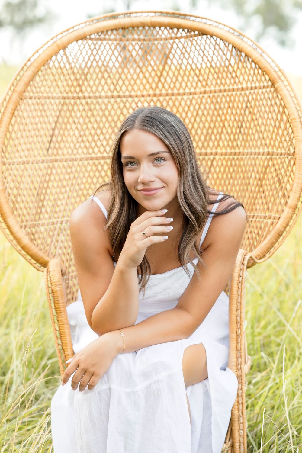 A person in a white dress poses on a vintage peacock chair in a sunlit field with tall grass on family's land in Ventura Iowa.