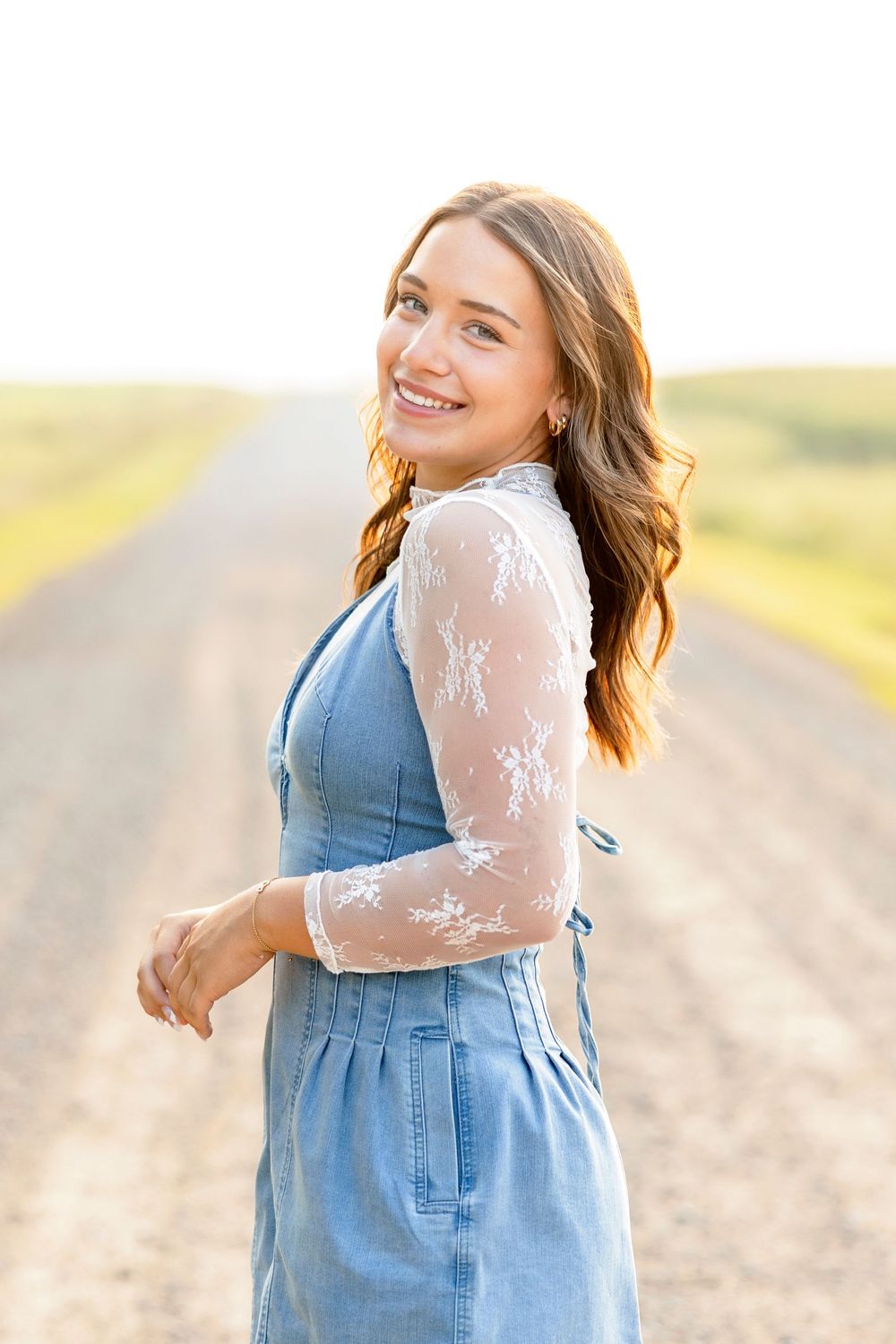 High School girl wearing blue denim dress and white lace top poses on a country road at sunset at family land in Clear Lake.
