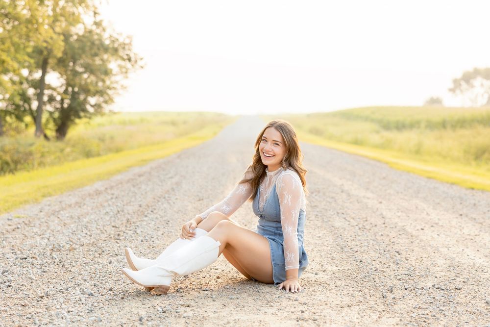 A person in a white dress and denim jacket poses on a rural gravel road during golden hour in North Iowa.