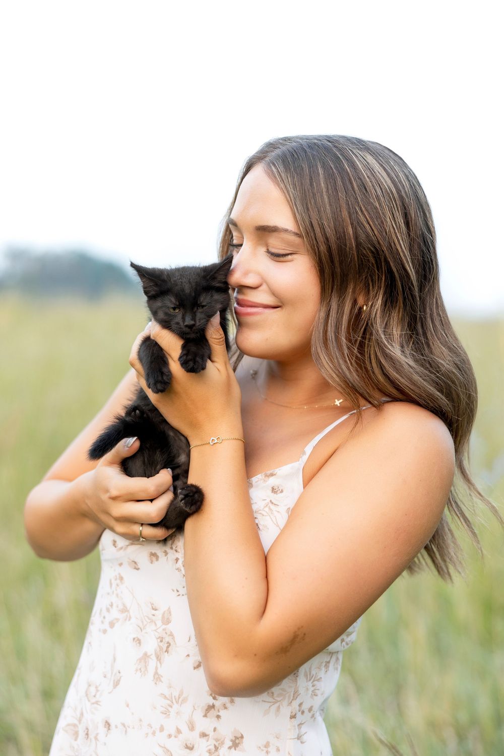 A person in a white lace dress holds and nuzzles a small black kitten in a sunny outdoor field during photo shoot.