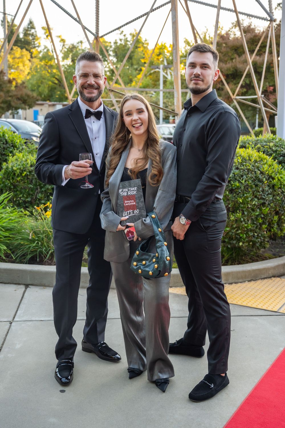 A group in formal attire laughs and poses together on a red carpet at an outdoor evening event.