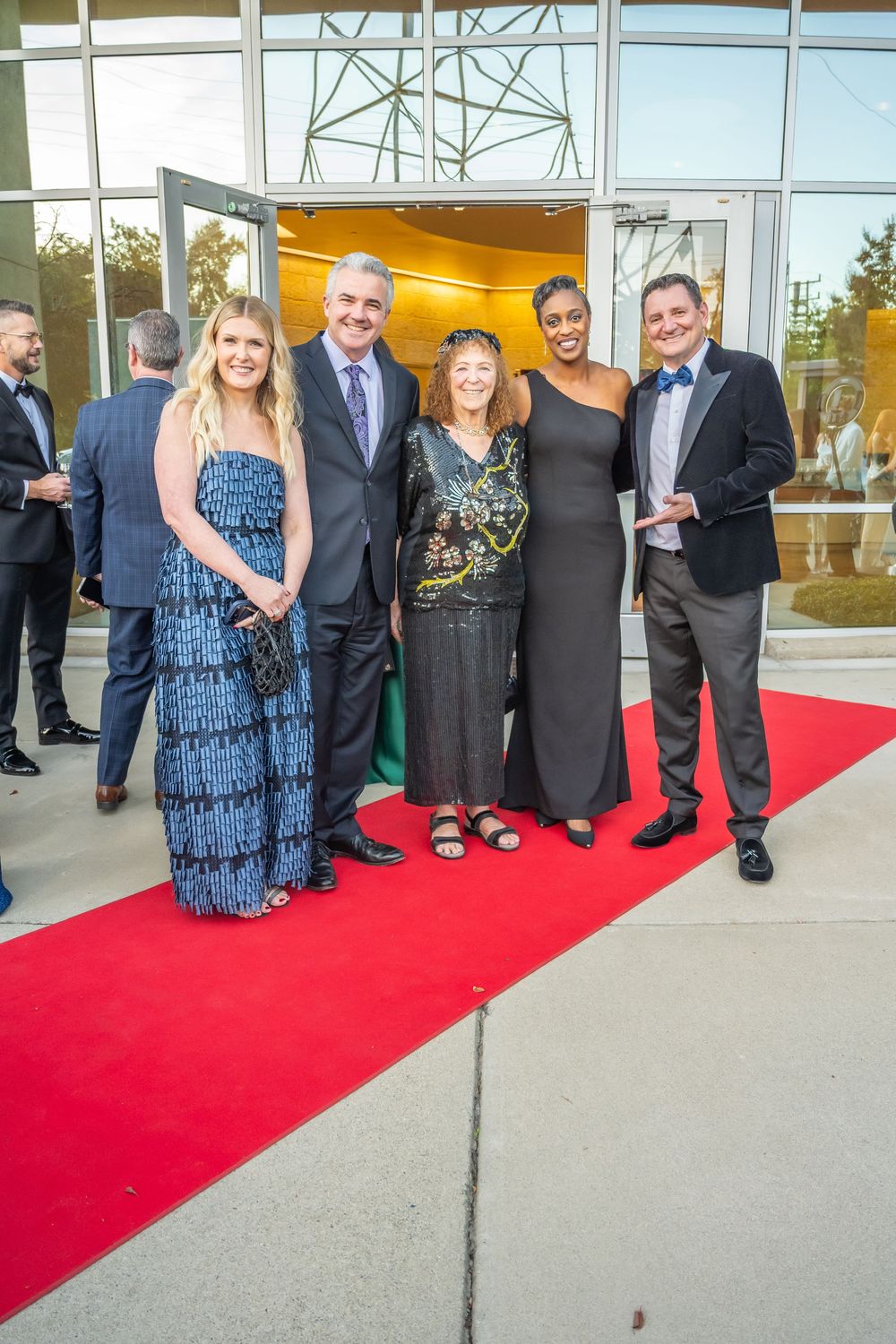 Group of formally dressed people posing together on a red carpet outside a modern glass building entrance.