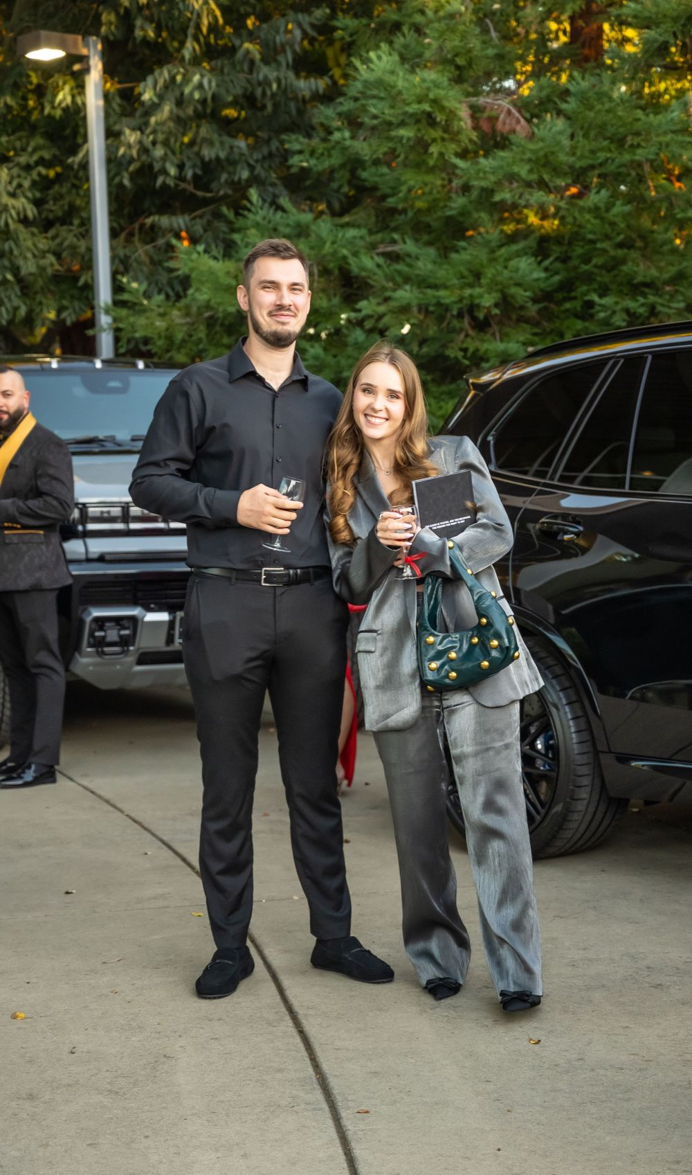 People in formal attire mingling in a parking lot area at dusk with cars in the background.