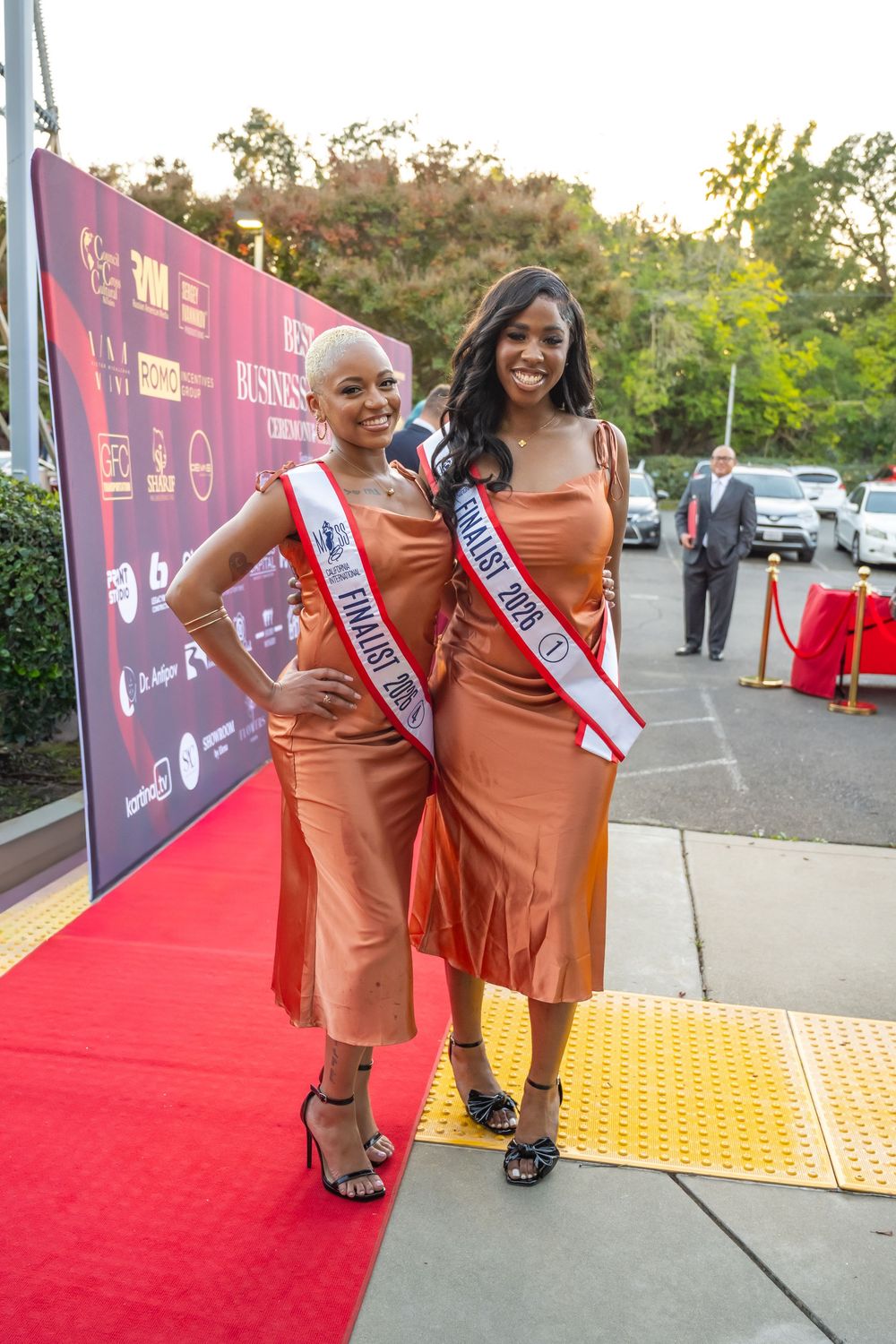 Attendees in matching orange dresses pose on a red carpet at an awards ceremony.