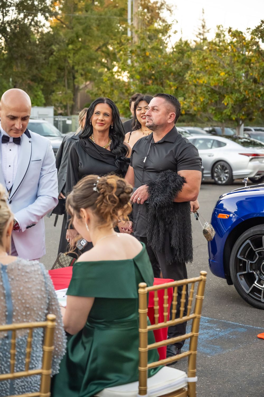 A group of people gather at an outdoor event with parked cars and gold chairs visible in the background.