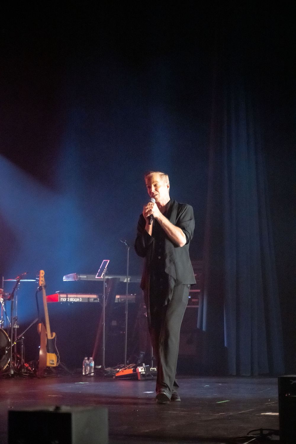 Performer on stage in dark lighting with musical equipment and instruments visible in the background.