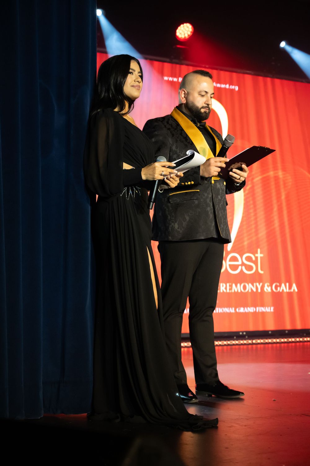 Presenters in formal attire reading from tablets on stage with dramatic lighting and red backdrop.