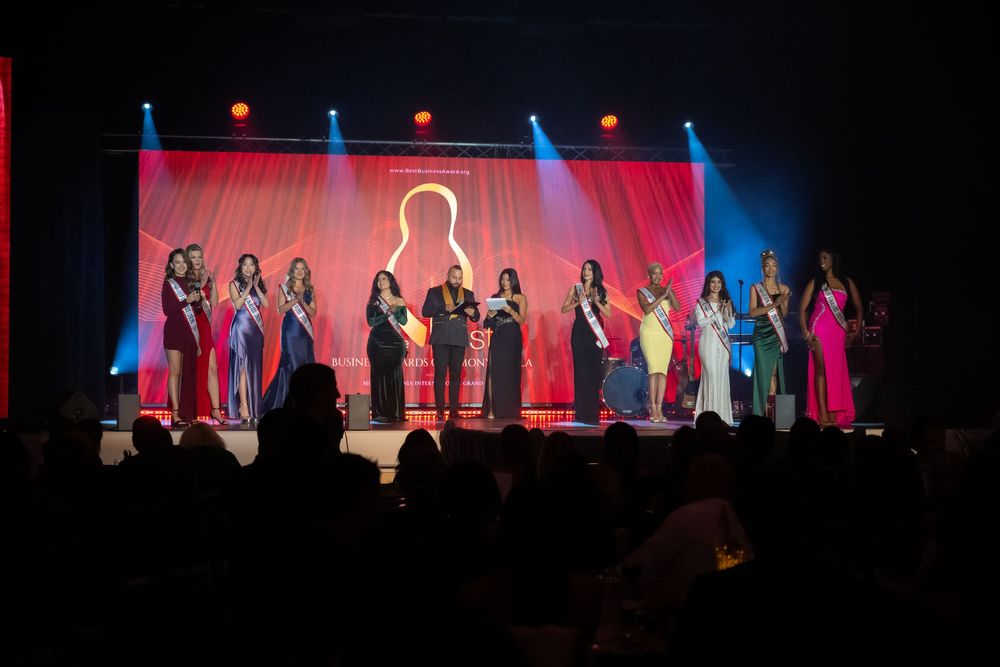 Beauty pageant contestants stand on stage in evening gowns under dramatic red and blue lighting during a competition.