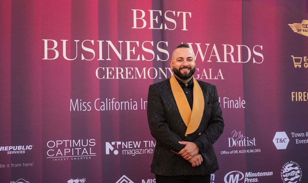 People in formal attire pose at a red carpet event with business awards ceremony backdrop and banners.